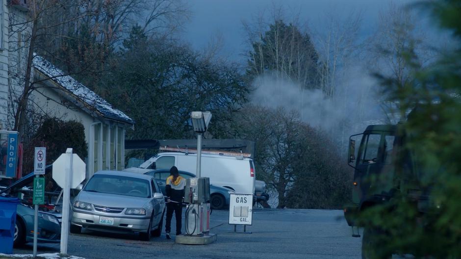 A man stops to pump gas as another military truck drives past.