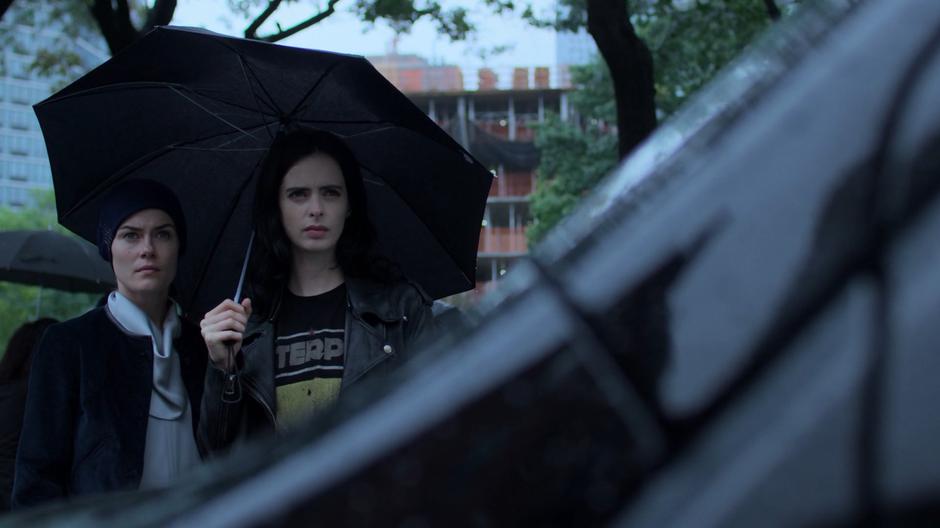 Trish and Jessica stand underneath an umbrella and watch the station from across the street.