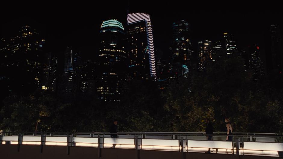 Bernard leads Dolores across a pedestrian bridge at night as a bunch of skyscrapers rise up in the background.