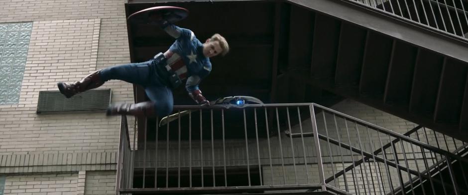 Steve leaps over the railing of a fire escape while holding Loki's staff.