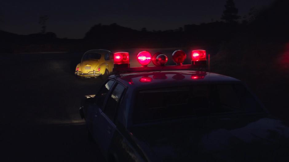 Bumblebee turns the corner in the dark while being followed closely by the police car.