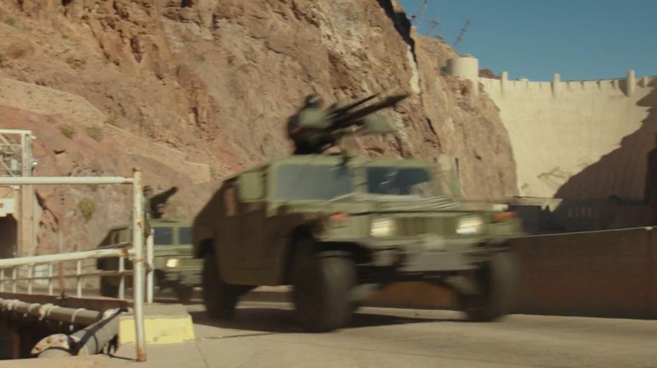 The mulitary Humvees drive across the bridge out of the base with the dam in the background.