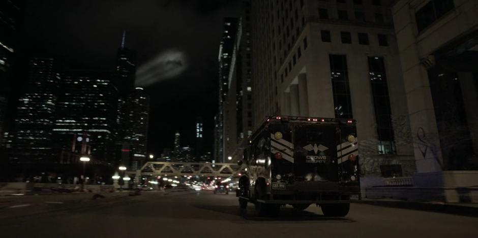 An armored Crows Security truck drives down the street towards where the Bat-Signal is shining in the sky.