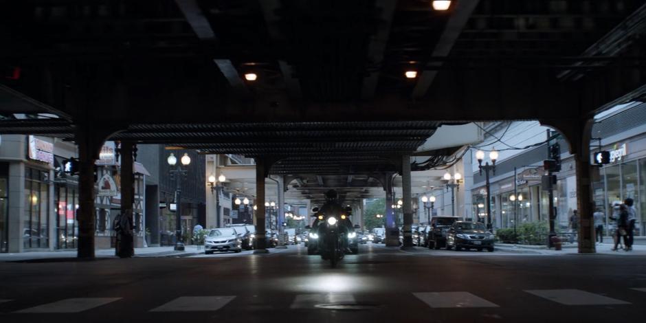 Kate rides her motorcycle down the street under the elevated tracks.