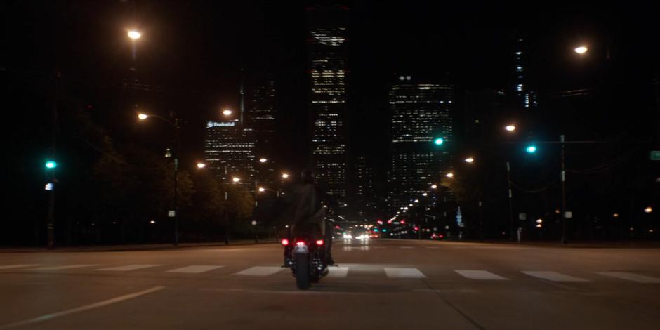 Kate rides her motorcycle through an intersection at night while wearing the Batsuit.