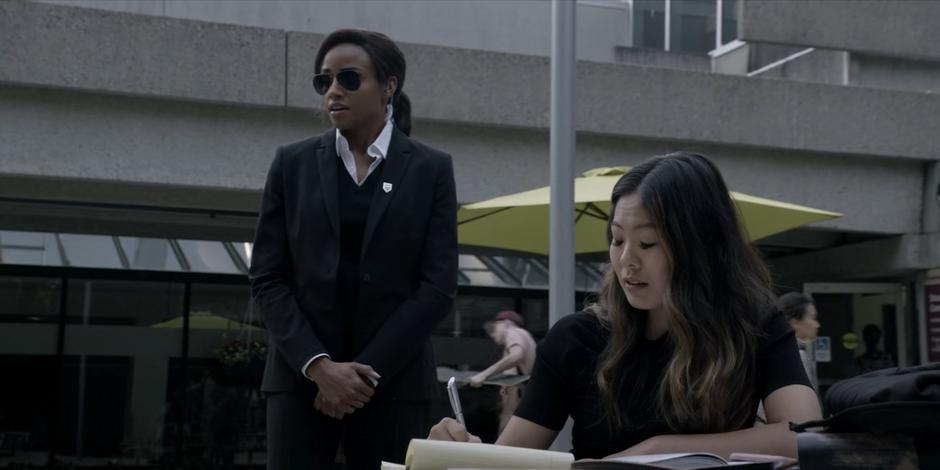 Sophie stands behind Mary and watches people walking past as Mary tries to study.