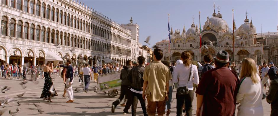 The tour group walks into the busy square as pigeons fly overhead.