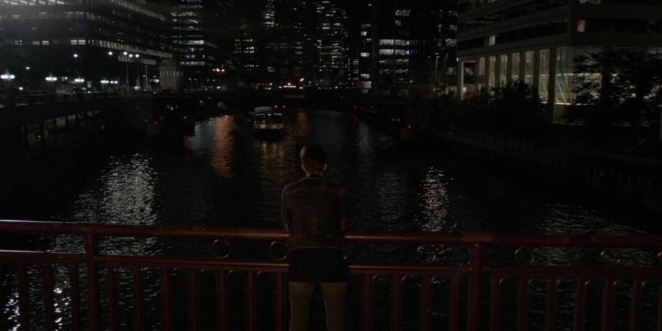 Kate leans on the bridge railing and looks out over the river at night.
