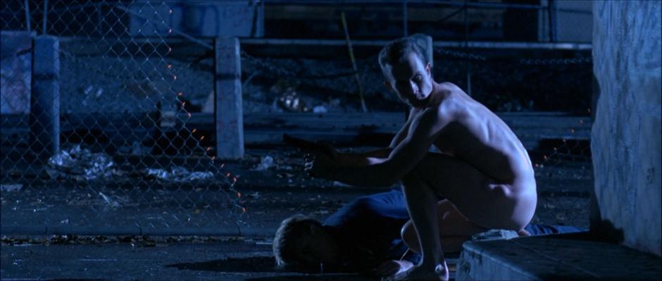 The naked T-1000 kneels over the body of the police officer and looks around while holding the officer's gun.