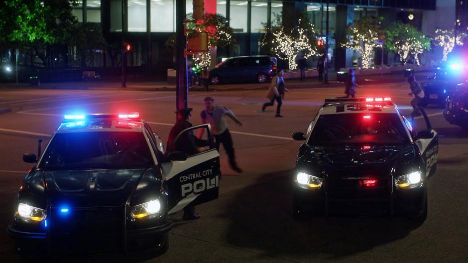 Police officers exit their cars as people run from the blood zombies.