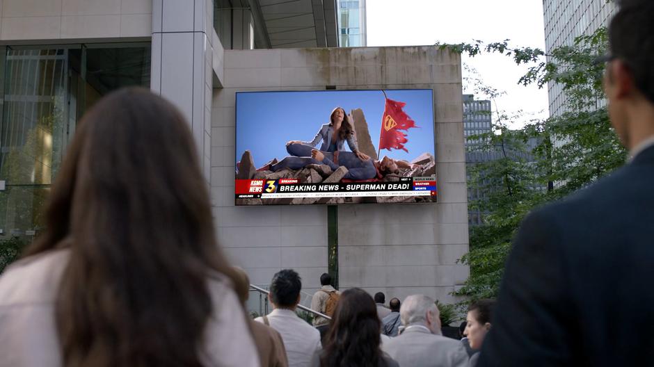 Lois and Clark look up at a screen showing a news report on the murder of this Earth's Superman by Lex.