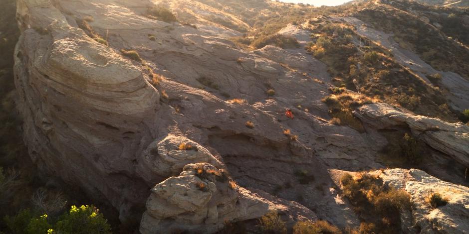 Tracy climbs up a steep rock face in her orange jumpsuit.