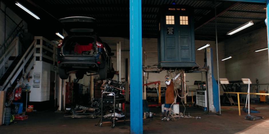 The Doctor works on the TARDIS which is elevated on a car lift.
