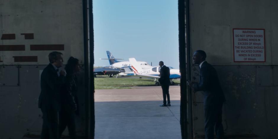 Graham, Yaz, and Ryan discuss what to do in whispers while hiding behind the hanger doors where Daniel Barton is standing outside.