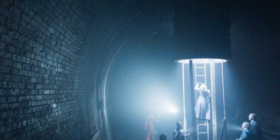 The Doctor climbs down the ladder into the tunnel while the others look around.