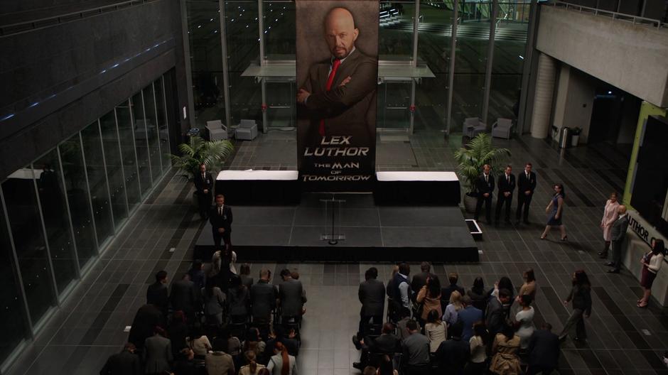 Lena walks to the lecturn for the press conference in front of Lex's giant photograph.