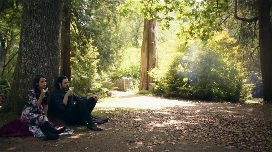 Margo and Eliot sit against a tree sipping cocktails.