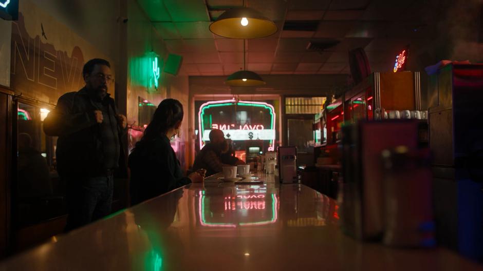 Ray adjusts his jacket as he prepares to head back into the alley. Mel looks back at him from the counter.