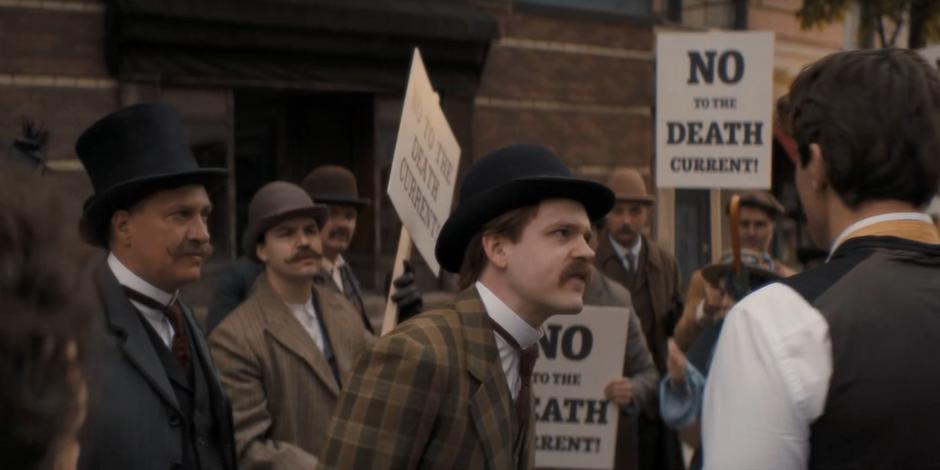 Nikola Tesla walks through the group of protestors in front of his lab.