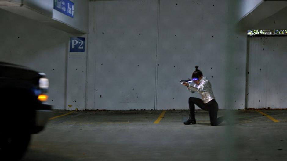 Kimiyo Hoshi kneels after dodging out of the path of Iris's car and raises her rifle to shoot again.