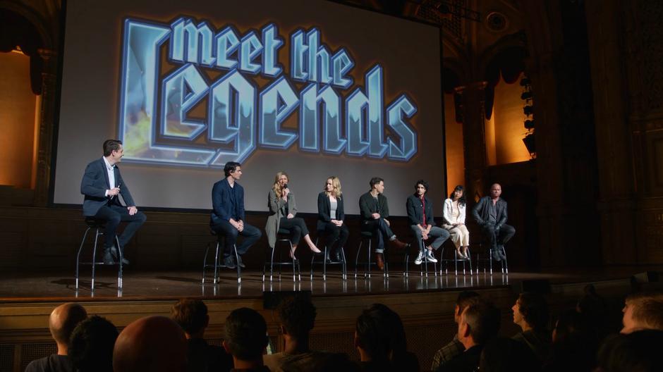 Kevin Harris watches as Ava torpedoes the Legends reputation. Ray sits to her left and Sara, Nate, Behrad, Mona, and Mick to her right.