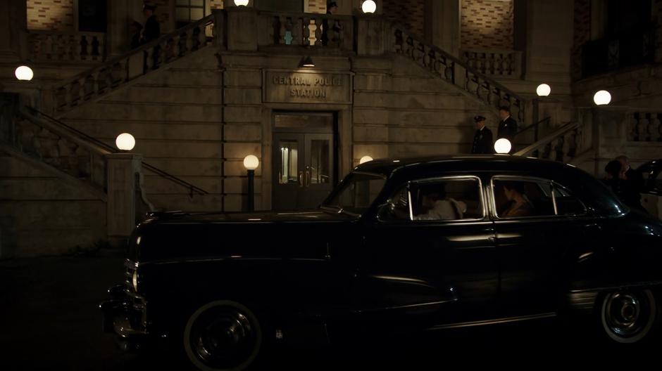 Constantine poses as Bugsy's driver and waits outside the police station for him with Jeanie Hill.