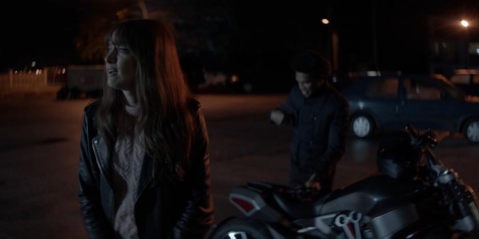 Beth looks around the empty parking lot while Luke stows his helmet on the Batcycle.