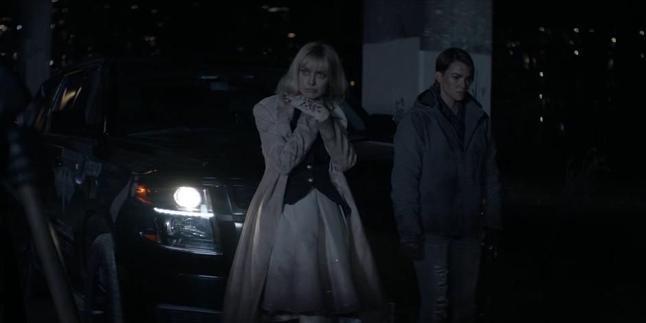 Alice mimes choking while Kate leans against the car next to her.
