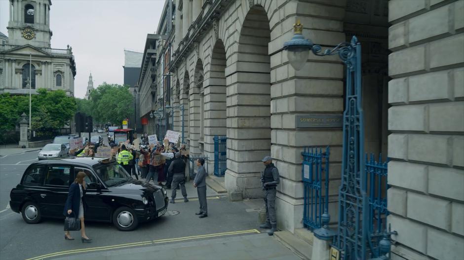 Myfanwy gets out of the taxi and walks to the gate as the police hold back the protestors.