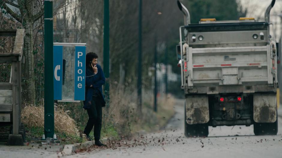 Jordan watches a dump-truck drive past as he talks on the payphone.