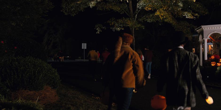 A bunch of trick-or-treaters walk down the street into the neighborhood.