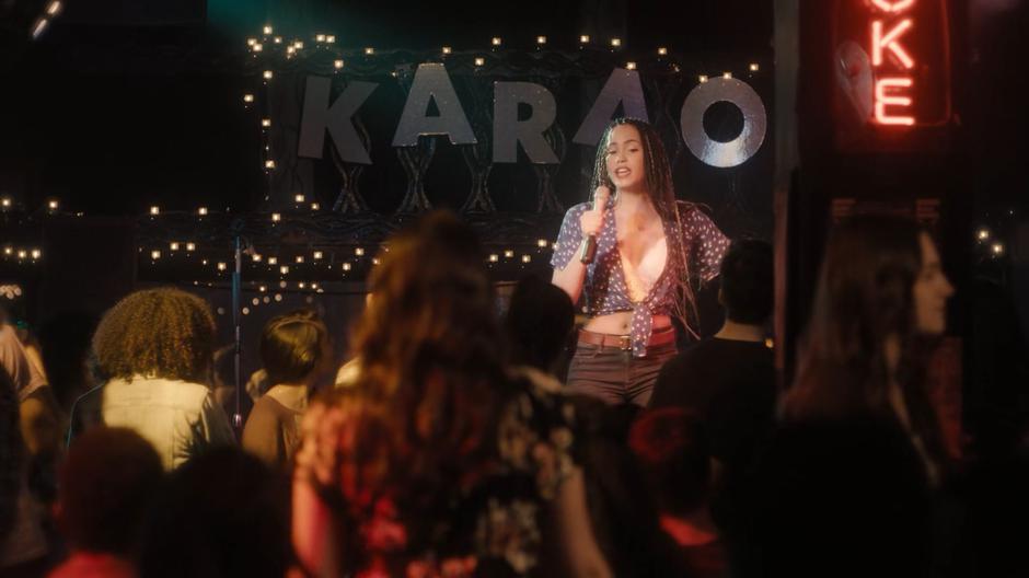 College-aged Macy sings karaoke on stage in a busy bar.