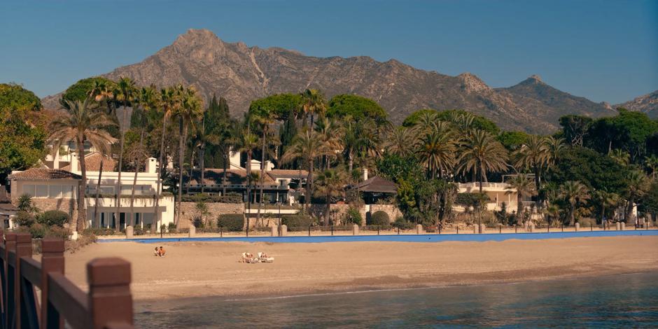 Ava and JC talk on the beach in front of the villa while the mountains rise behind them.