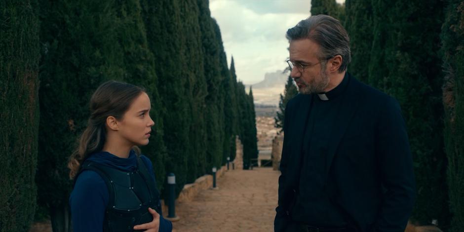 Ava stops to talk with Father Vincent as they walk up the path to the training ground.