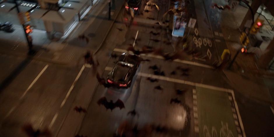 The Batmobile speeds through an intersection as the swarm of bats fly overhead.