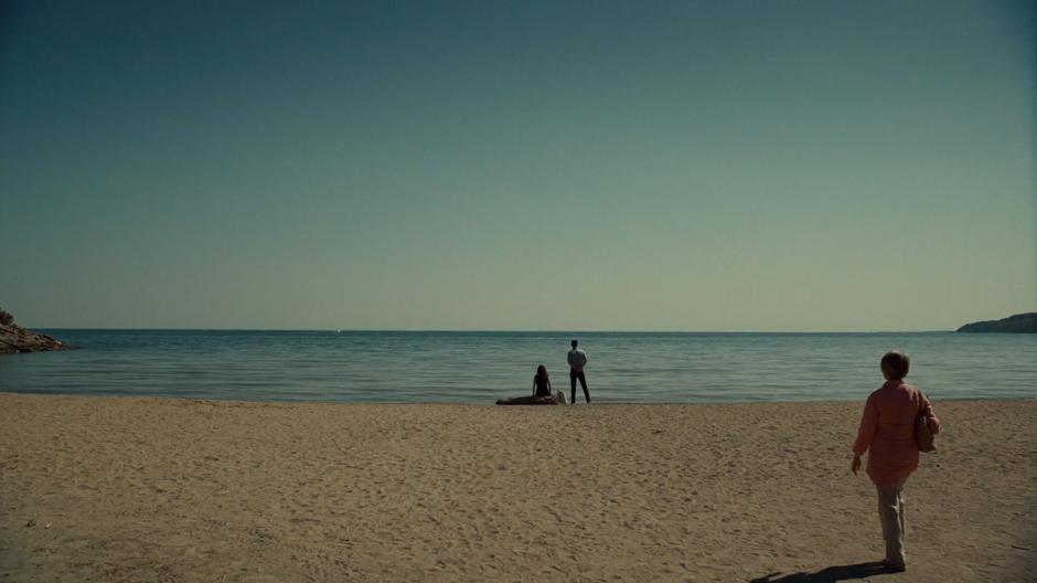 A woman approaches Mary and Des who are waiting on the beach.