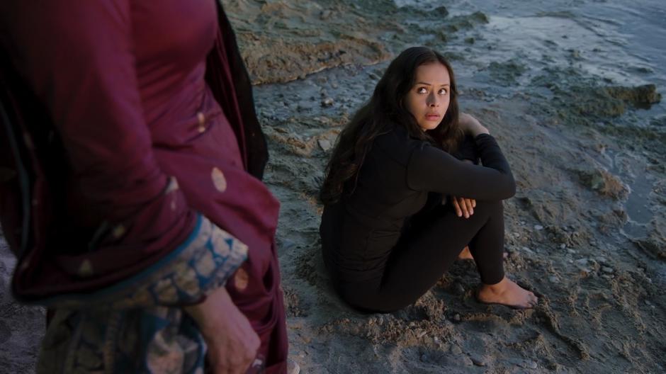 Bobby looks back over her shoulder from where she is sitting on the sand as Chrisjen approaches.