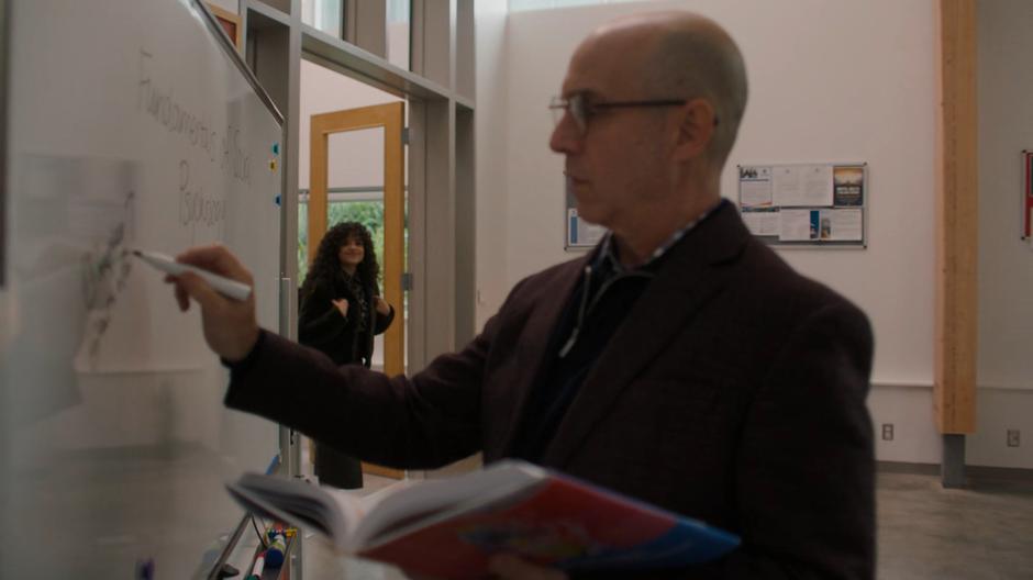 Maggie enters the classroom and sees her returning professor Dr. Lawrence writing on the whiteboard.