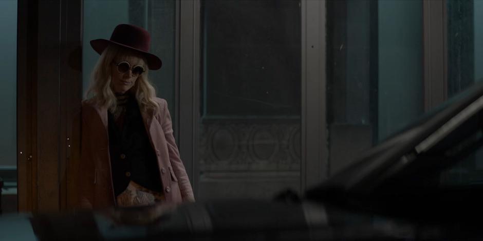 Alice waits for Tatiana on the side of the street in a fancy hat.