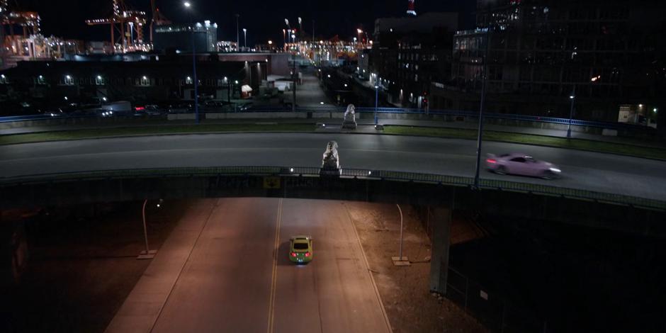 The green car continues under the overpass while the pink one drives overhead.