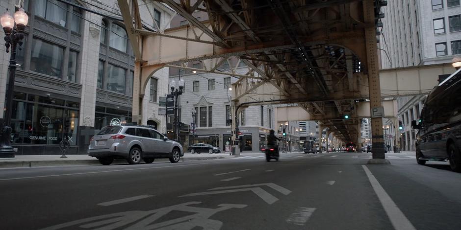 Kate speeds down the street beneath the elevated tracks on her motorcycle.