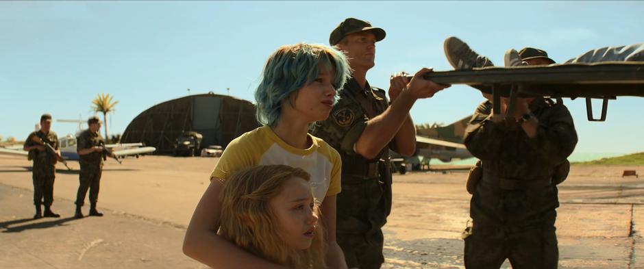 Natasha and Yelena watch as soldiers load Melina into the back of a truck.