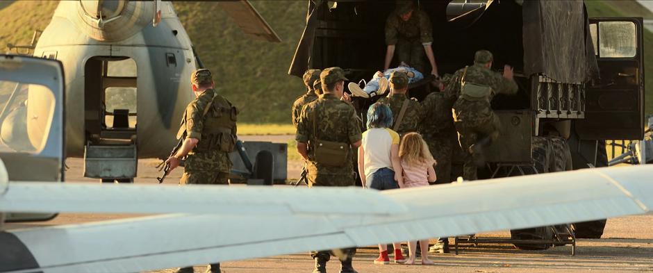 Soldiers load Melina into the back of a truck on a stretcher while Natasha and Yelena watch.