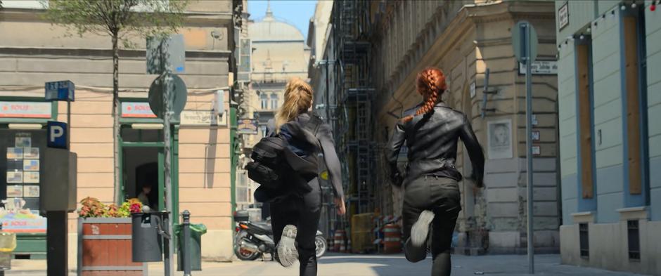 Yelena and Natasha run down the street towards some parked motorcycles.