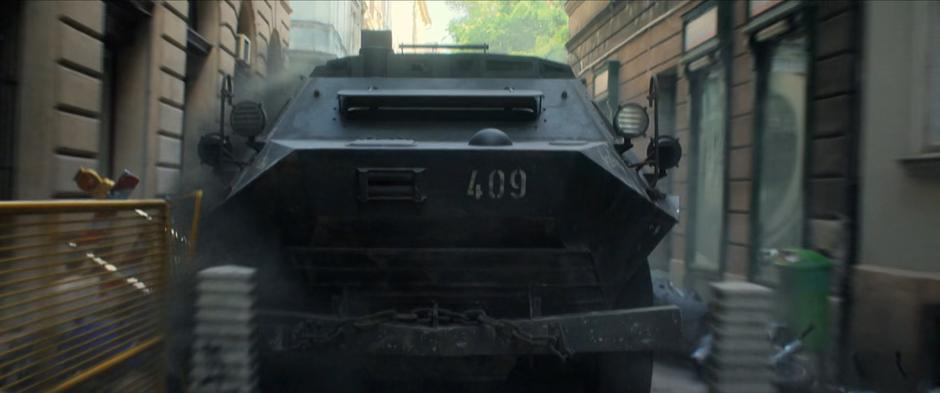 The armored vehicle plows its way down the alley through barriers.