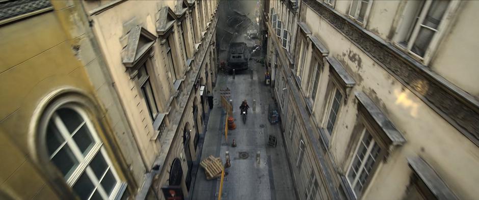 Natasha and Yelena speed down the alley just barely in front of the armored vehicle.