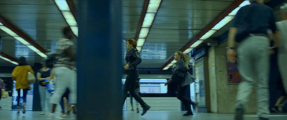 Natasha and Yelena run across the busy platform.