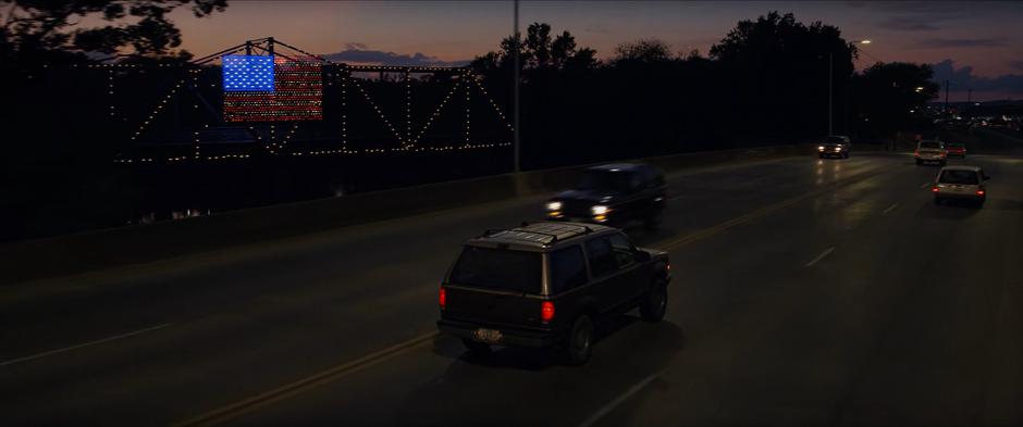 The family drives past a rail bridge with an American flag made of lights hanging from it.