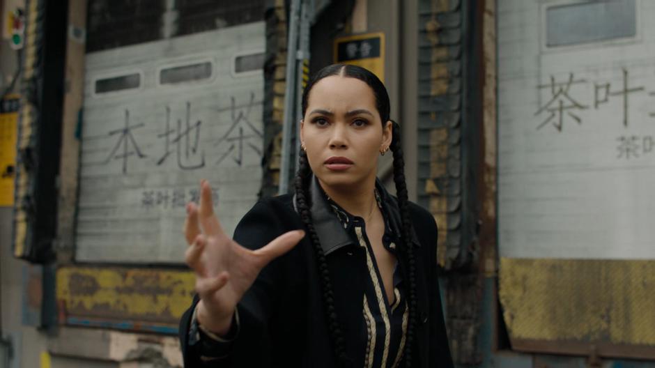 Macy appears in front of a loading dock with her arm outstretched after being teleported away.