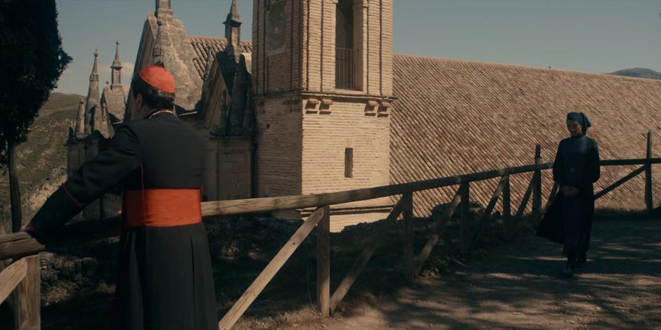 Lilith approaches Cardinal Duretti as he is standing on the path above the building.